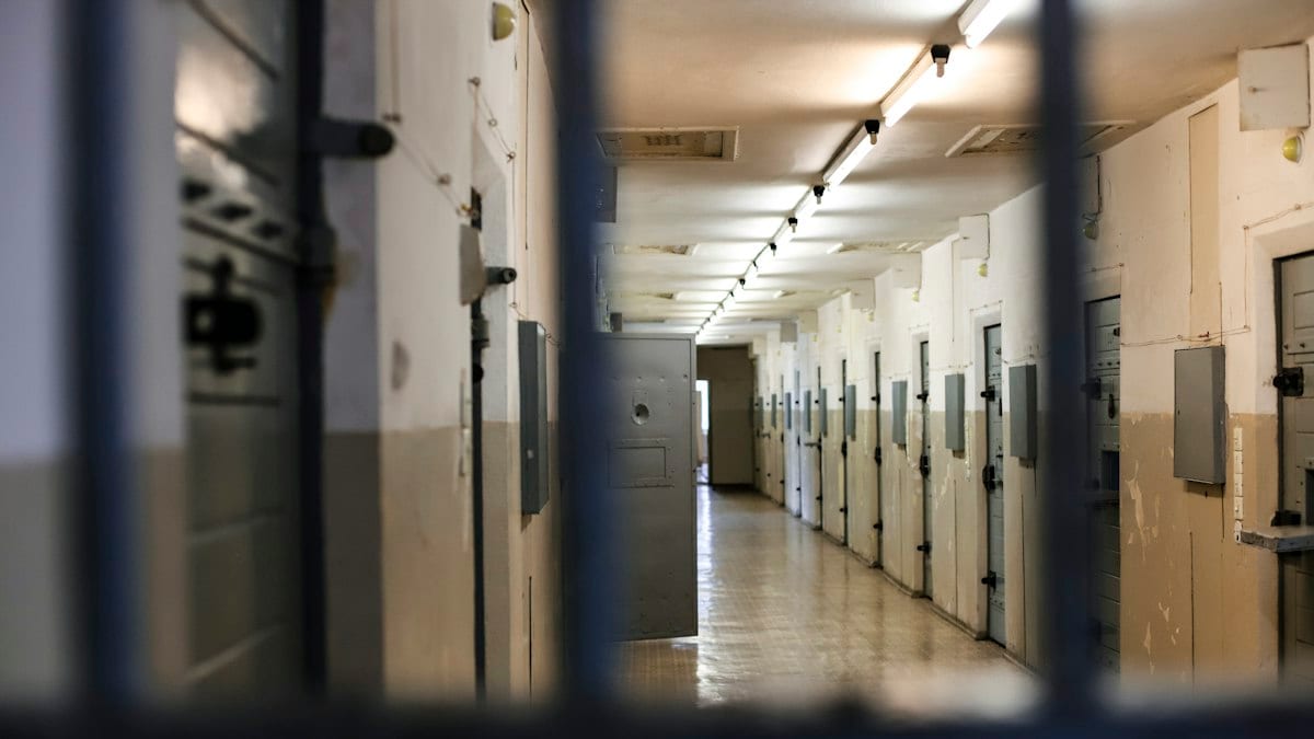 A long prison corridor with rows of metal doors viewed through dark bars