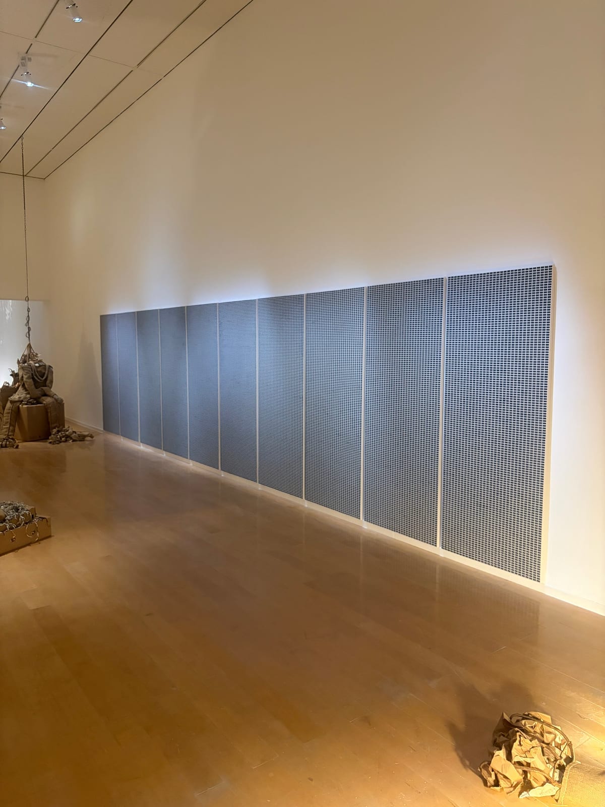 Un long mur de panneaux couverts d'une grille dense de petites cases bleu-gris, dans une salle de musée au parquet clair