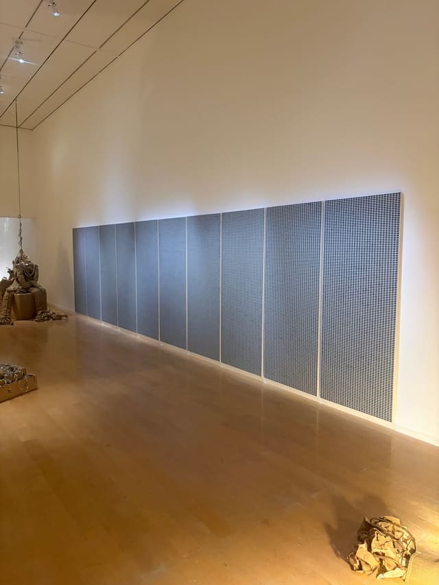 Un long mur de panneaux couverts d'une grille dense de petites cases bleu-gris, dans une salle de musée au parquet clair