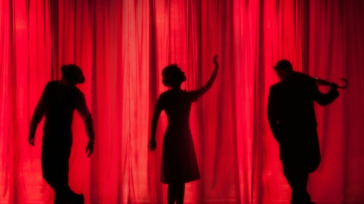 Trois silhouettes d'acteurs se détachent devant un rideau de scène rouge.