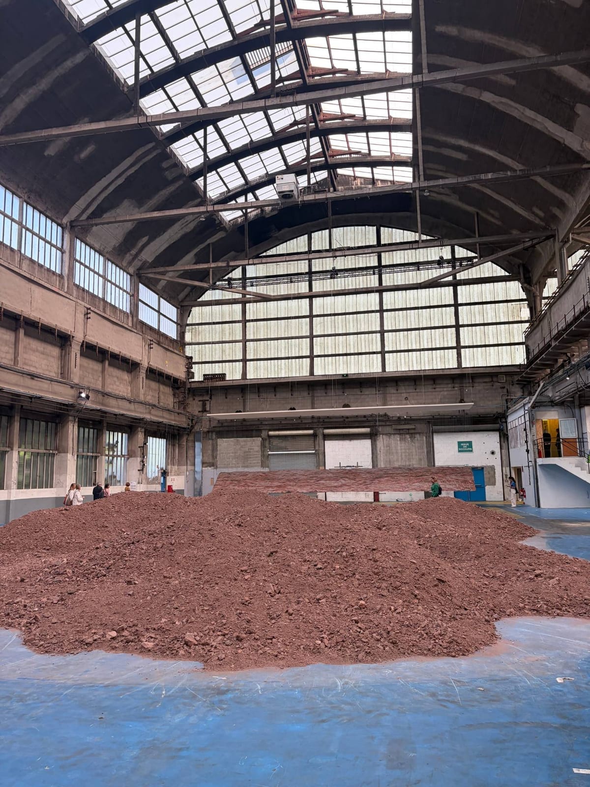 Une dune de schiste rouge occupe le sol d'une vaste halle industrielle à verrière ; quelques visiteurs à ses pieds en donnent l'échelle