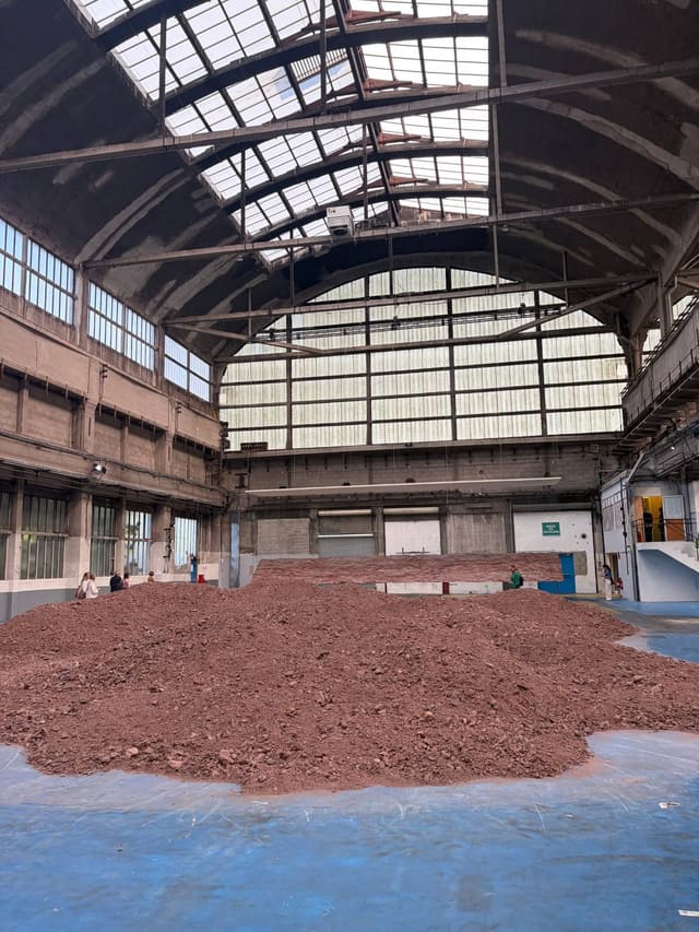 Une dune de schiste rouge occupe le sol d'une vaste halle industrielle à verrière ; quelques visiteurs à ses pieds en donnent l'échelle