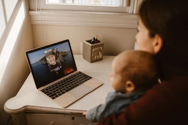 A mother holding her baby while making a video call on a laptop indoors.
