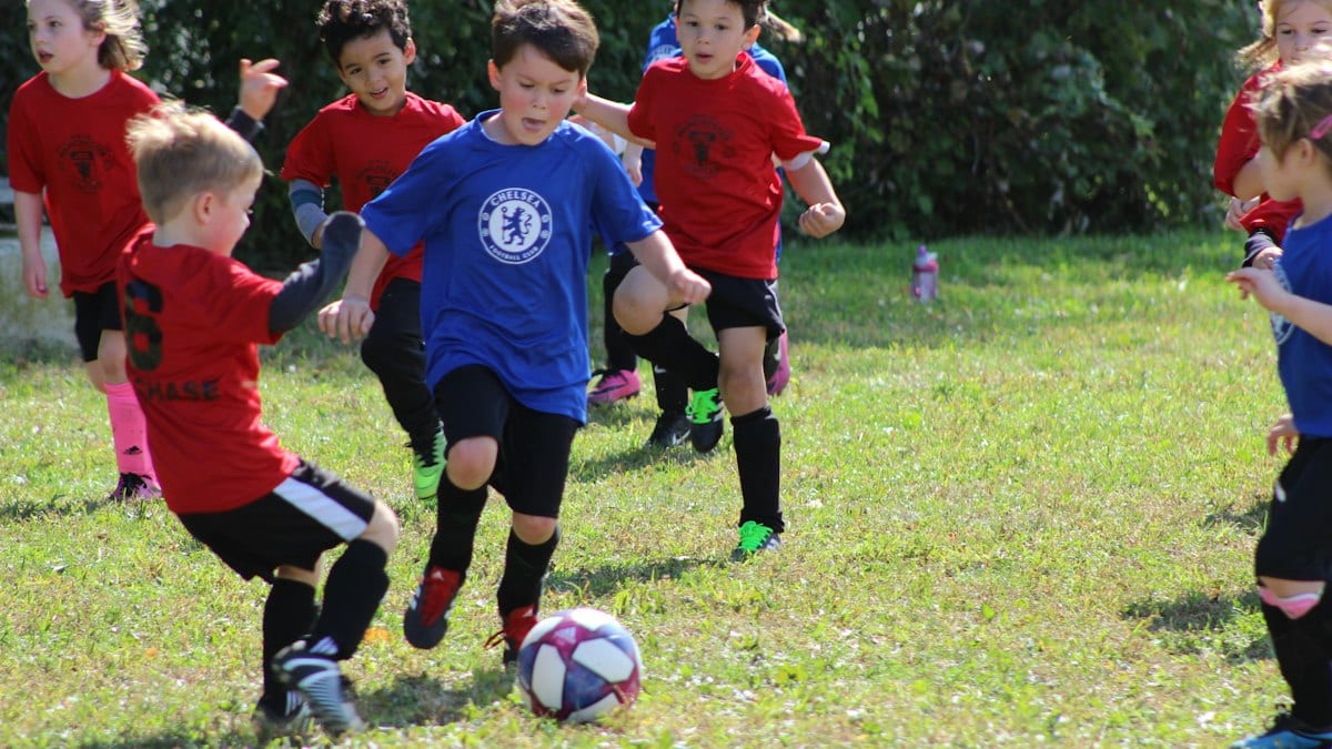 boy in red and blue soccer jersey kicking soccer ball on green grass field during daytime