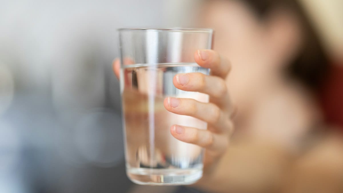 Une main tend un verre d'eau transparent devant un visage flou.