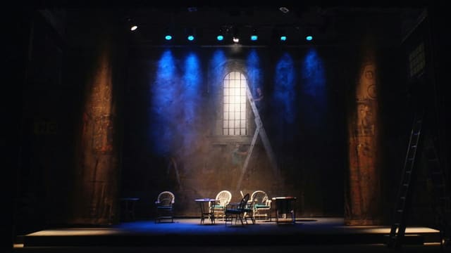 Scène de théâtre plongée dans la pénombre, éclairée de projecteurs bleus, avec quelques chaises et une échelle appuyée contre un décor.