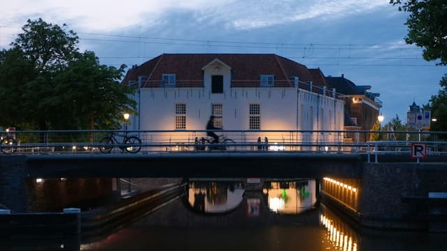 Un cycliste traverse un pont au crépuscule, devant une maison éclairée au bord d'un canal.