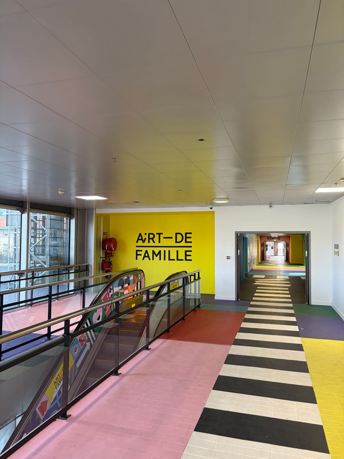 Une passerelle intérieure aux sols peints en rose, jaune et violet mène à un escalier mécanique ; sur le mur du fond, le logo AiRT—DE FAMILLE en noir sur jaune