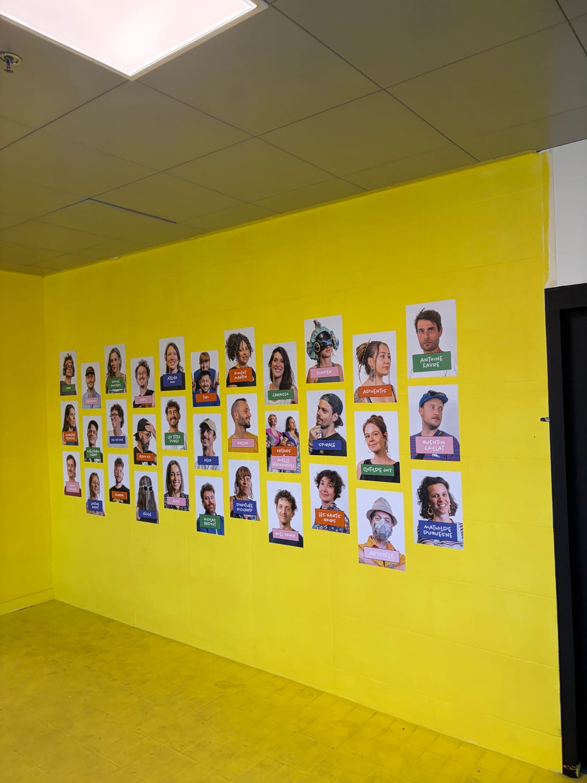 Un mur peint en jaune couvert d'une trentaine de portraits photographiques d'artistes, chacun légendé d'un nom ou d'un pseudonyme sur une étiquette de couleur