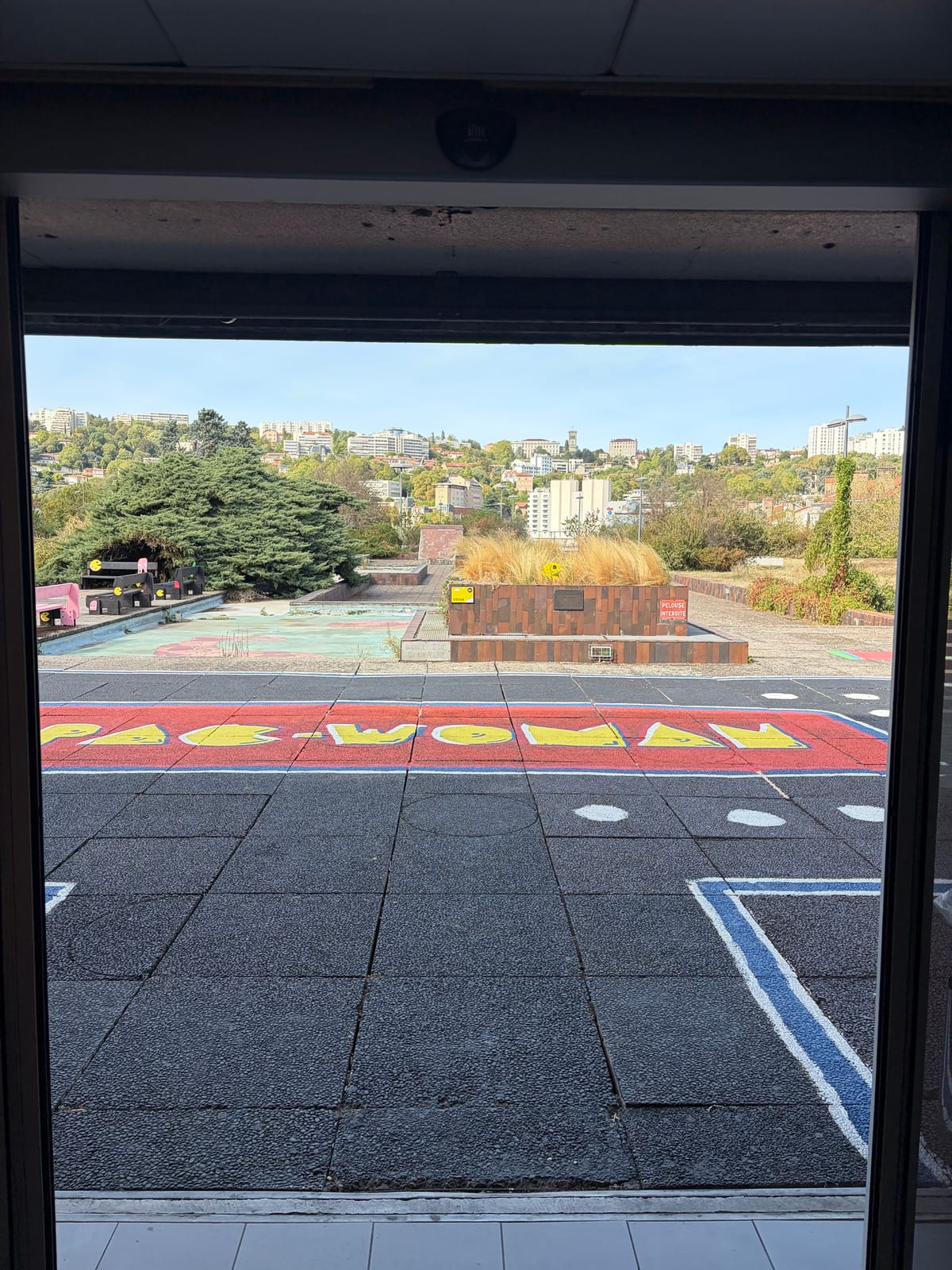 Vue depuis une baie vitrée sur une terrasse en dalles sombres où est peint en lettres jaunes « PAC-WOMAN », avec des bancs roses et, au fond, les collines de l'ouest lyonnais