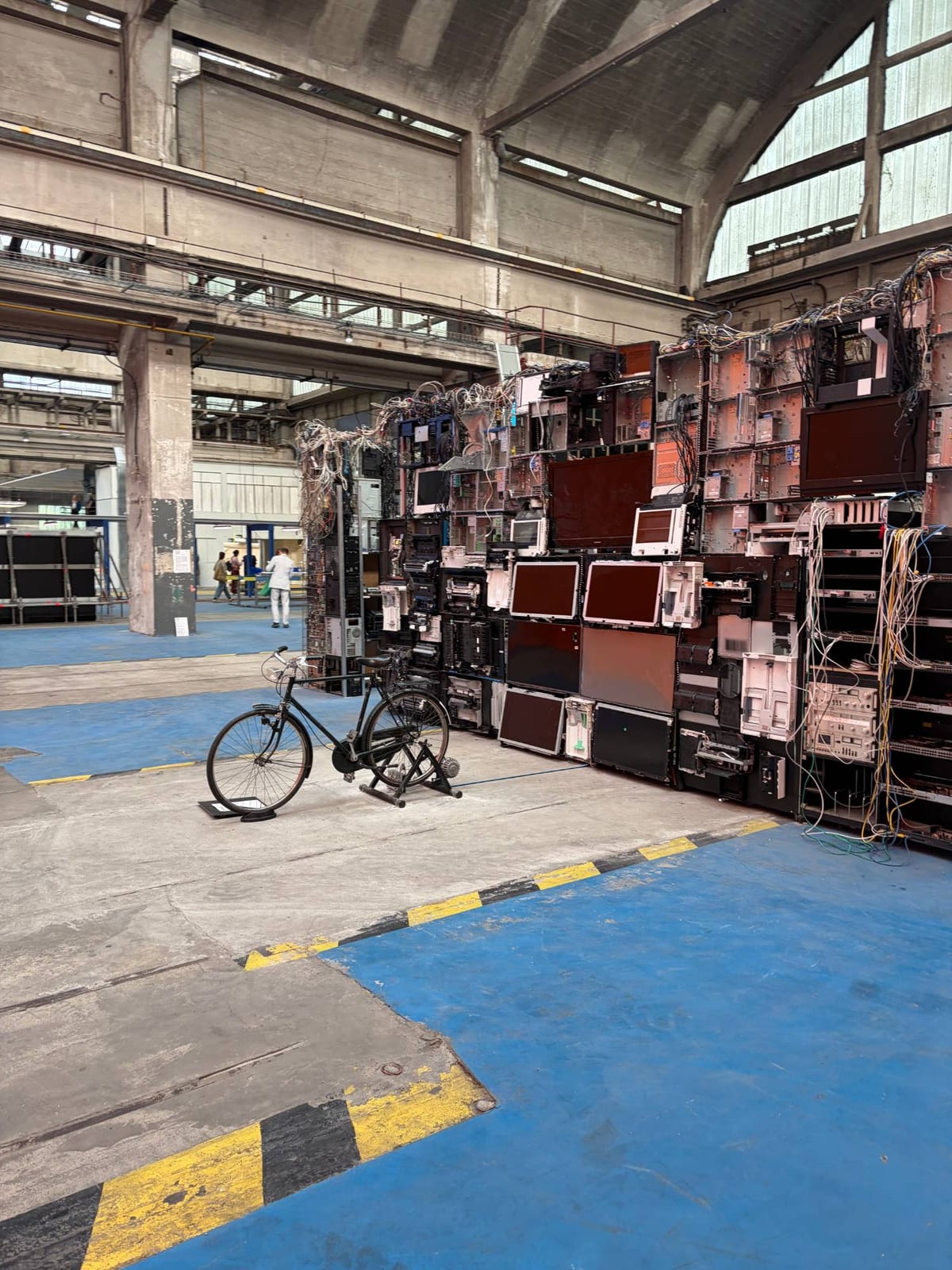 Un mur monumental d'écrans démontés, d'unités centrales éventrées et de faisceaux de câbles dressé dans la halle ; un vélo est posé devant, relié à l'installation