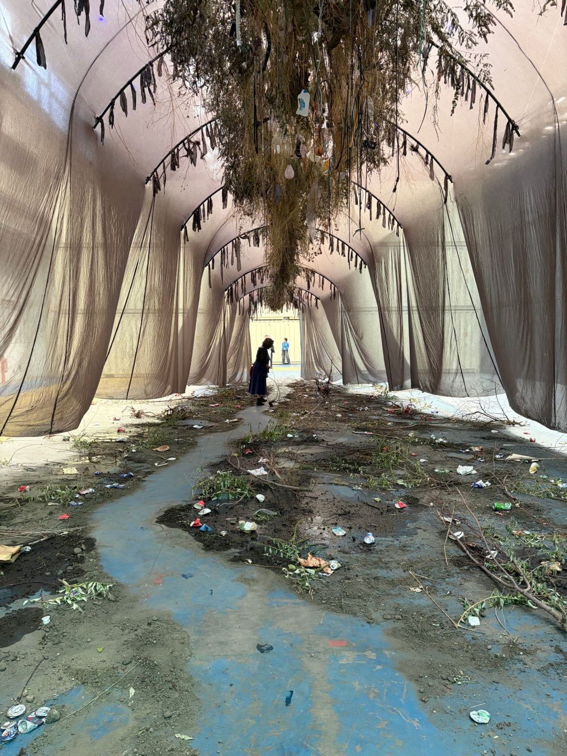 Sous une voûte de textiles tendus, un sol jonché de branchages, de canettes et de déchets plastiques traversé par une coulée bleue ; une visiteuse se tient au fond