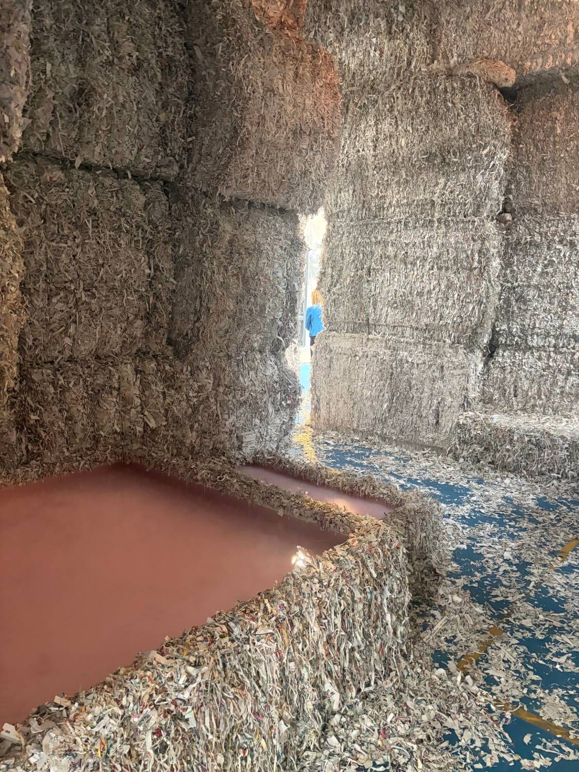 Un dédale de murs faits de balles de papier broyé et compressé, percé d'une ouverture étroite où passe une visiteuse ; au premier plan, un bassin d'eau rose