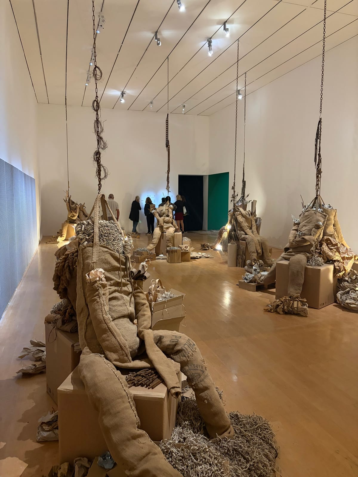 Plusieurs figures à taille humaine cousues dans des sacs de jute, affaissées sur des cartons et suspendues à des chaînes dans une grande salle blanche