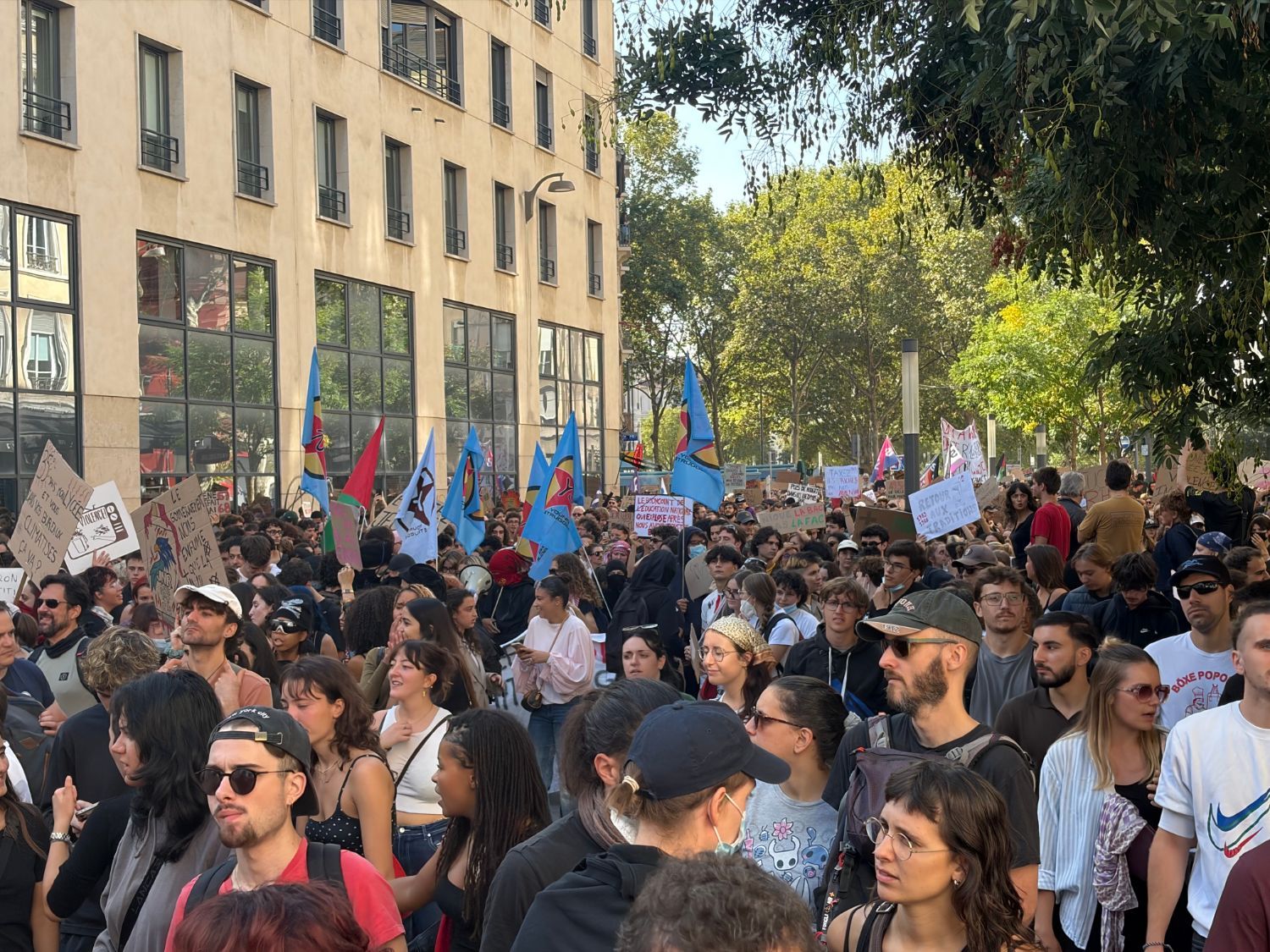 Le cortège avance dans une rue bordée d'arbres, drapeaux et pancartes au-dessus de la foule