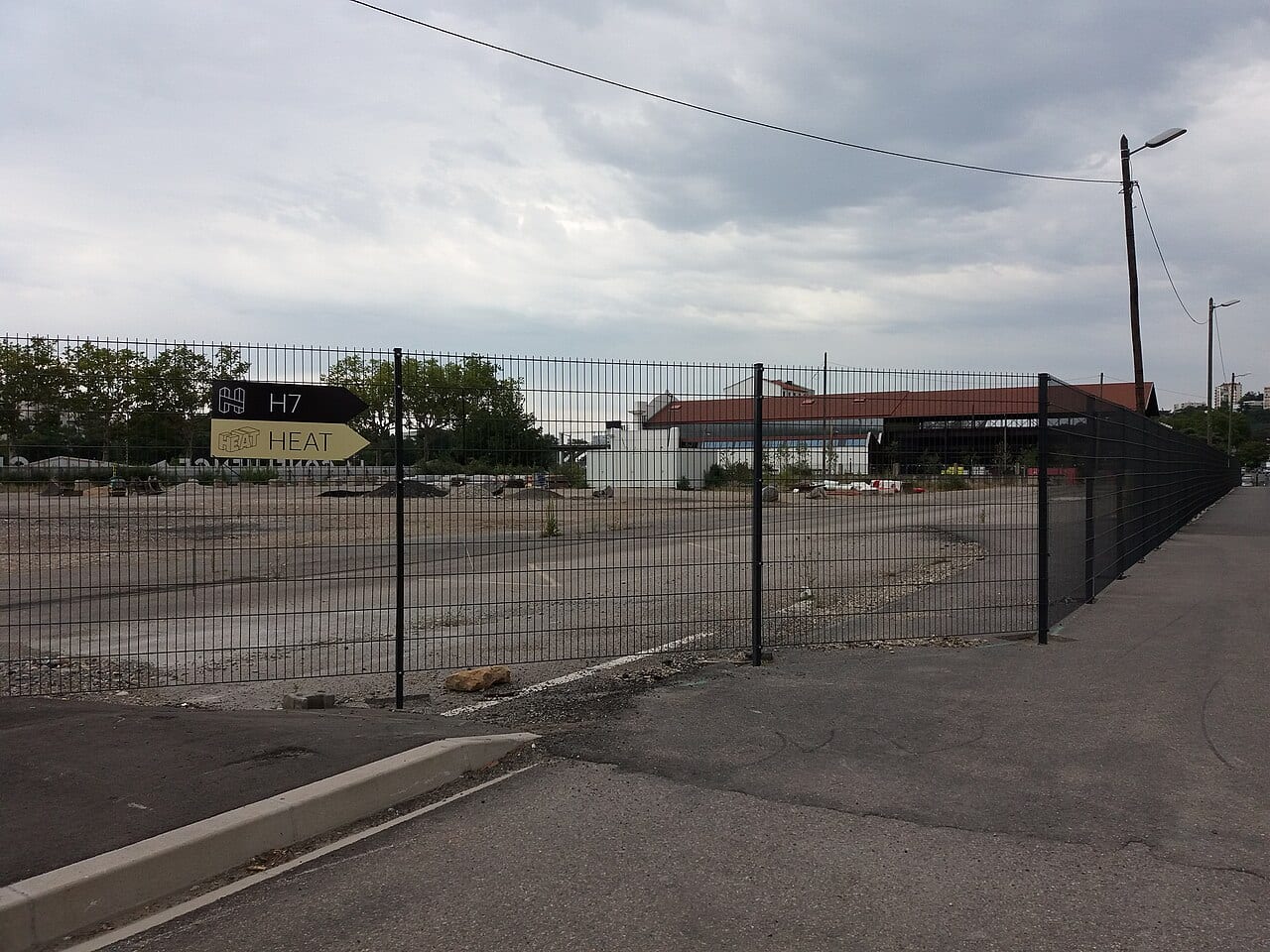 Panneau fléché vers le H7 derrière une clôture, avec la halle à toiture rouge en arrière-plan