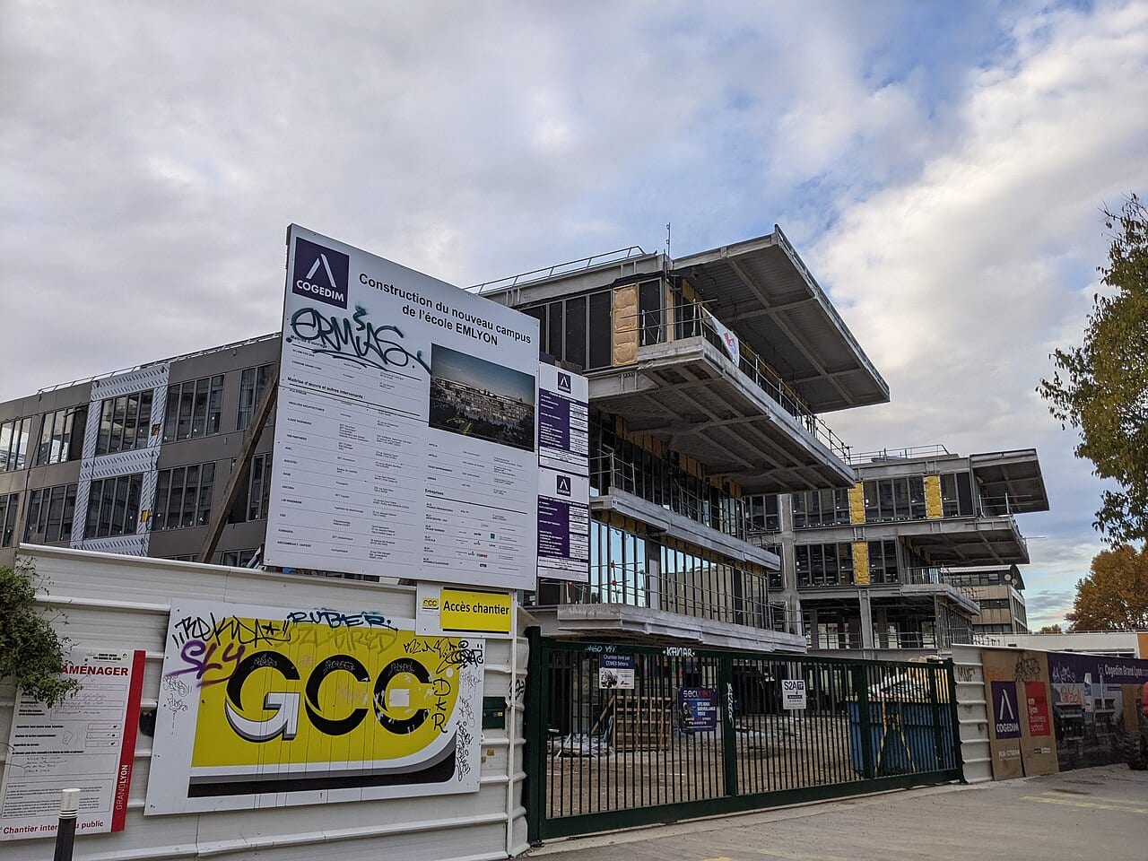 Bâtiments du campus emlyon business school pendant leur construction, avec panneau de chantier et palissade ornée de graffitis.