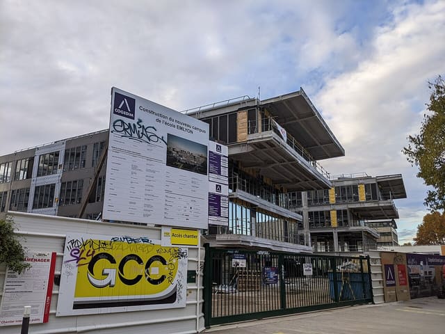 Bâtiments du campus emlyon business school pendant leur construction, avec panneau de chantier et palissade ornée de graffitis.