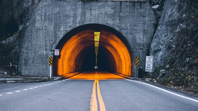 Entrée d'un tunnel routier éclairé d'orange, de nuit, au bout d'une route à double ligne jaune.