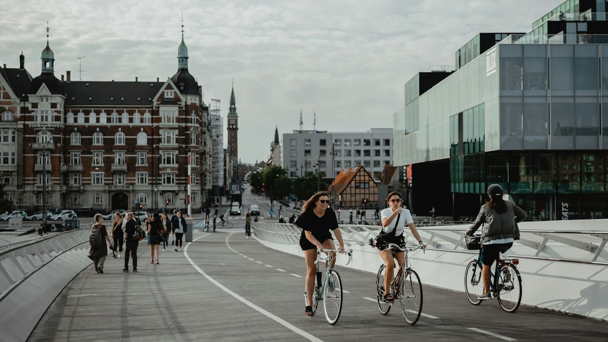 two people biking and other people walking on pathway near buildings under white and blue sky