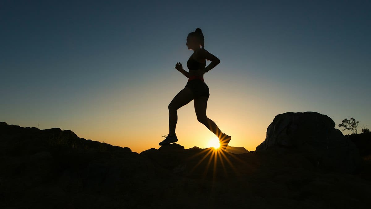 Silhouette d'une coureuse de trail en contre-jour, sur un sentier rocheux au coucher du soleil.