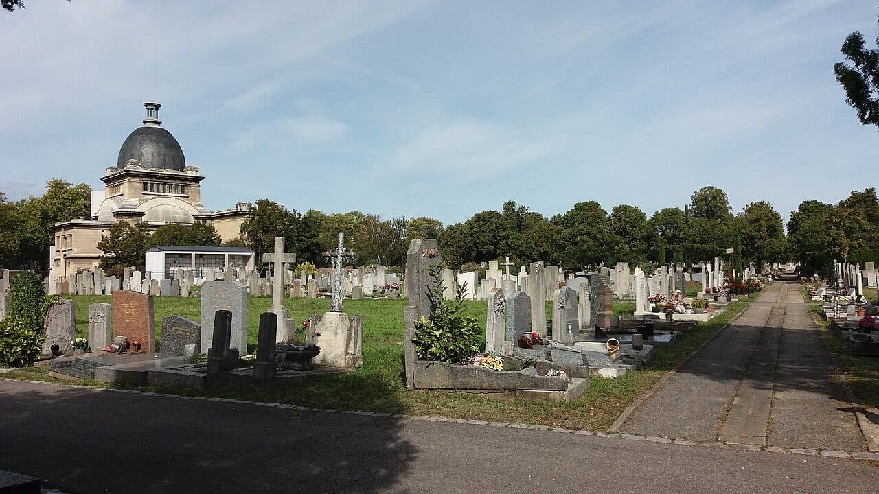 Allées et tombes d'un cimetière lyonnais sous un ciel dégagé, avec un bâtiment à dôme en arrière-plan