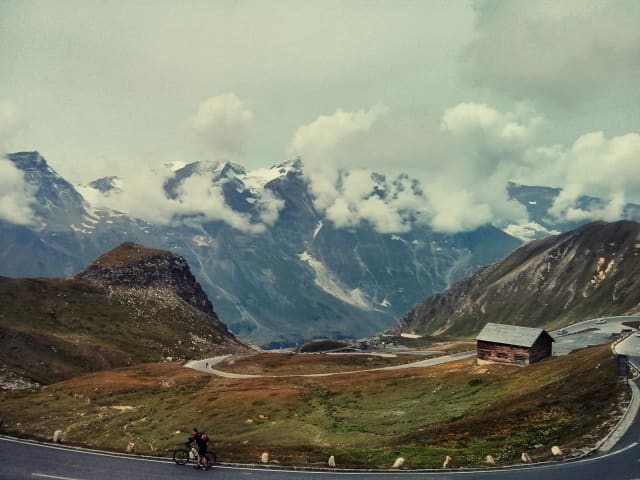 Picturesque view of a winding alpine road with a wooden hut and dramatic mountain backdrop.
