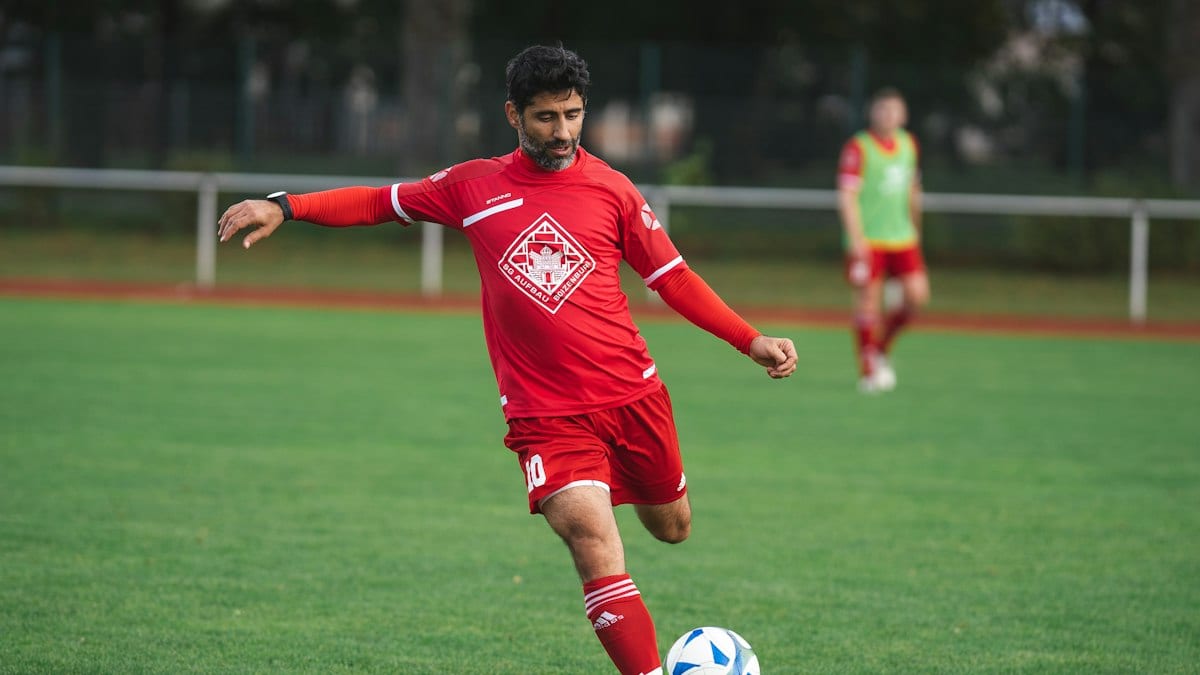 Un joueur en maillot rouge frappe le ballon sur une pelouse lors d'un entraînement de football amateur, un coéquipier en arrière-plan.