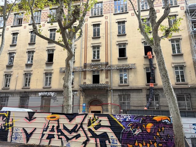 La façade du lycée La Martinière, portant l'inscription « La Martinière — École nationale professionnelle », vue depuis la rue derrière des platanes, palissade de chantier et échafaudages au pied du bâtiment
