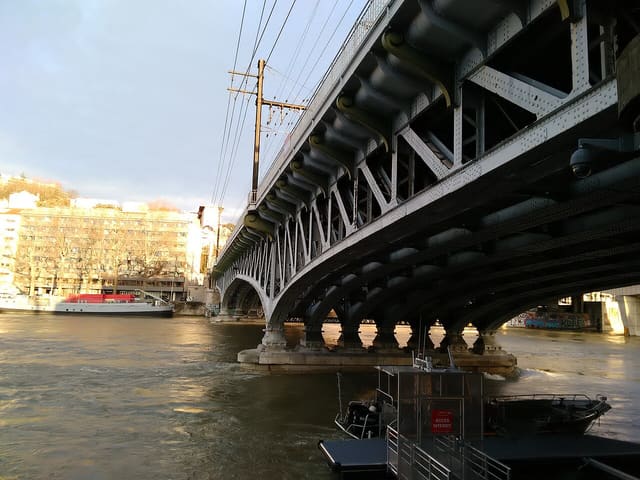 Un pont métallique à arches enjambe une rivière aux eaux brunes, avec un bateau amarré à l'arrière-plan et des immeubles sur la berge.