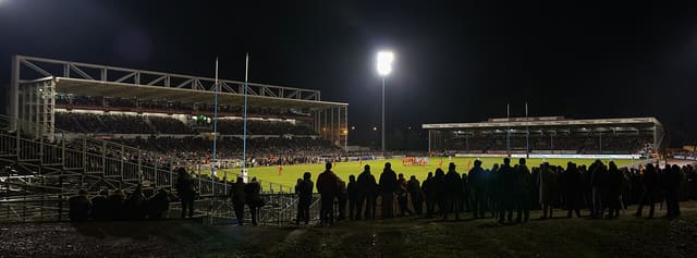 Vue de nuit d'un stade de rugby éclairé par un projecteur, tribunes garnies de spectateurs autour de la pelouse