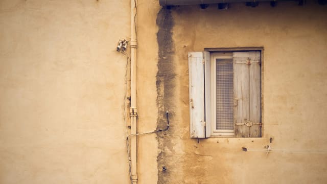 Façade ocre d'une maison ancienne avec une fenêtre aux volets blancs et une trace sombre le long du mur