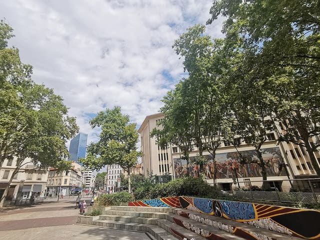 La place Guichard, dans le 3e arrondissement de Lyon, avec la Bourse du Travail et la fresque peinte sur sa façade, sous les platanes.