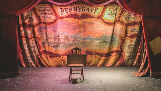 Scène de théâtre vide, éclairée en rouge, avec une chaise devant un grand rideau de fond peint aux motifs de foire ancienne.