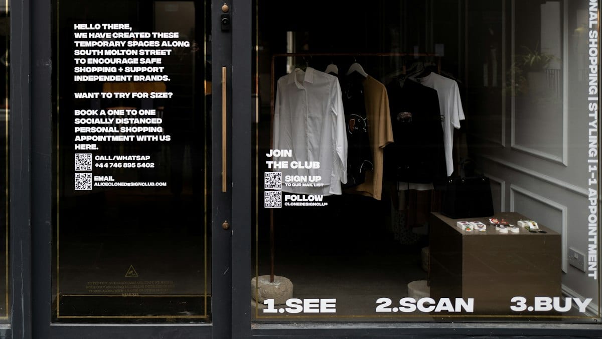 Vitrine d'une boutique de vêtements avec des affichages blancs sur la vitre, devant un portant de chemises.