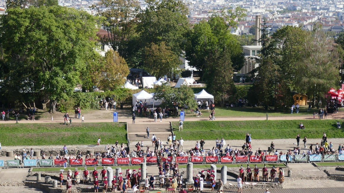 Public rassemblé en plein air sur le site archéologique de Fourvière, avec tentes blanches et verdure, et la ville de Lyon en arrière-plan