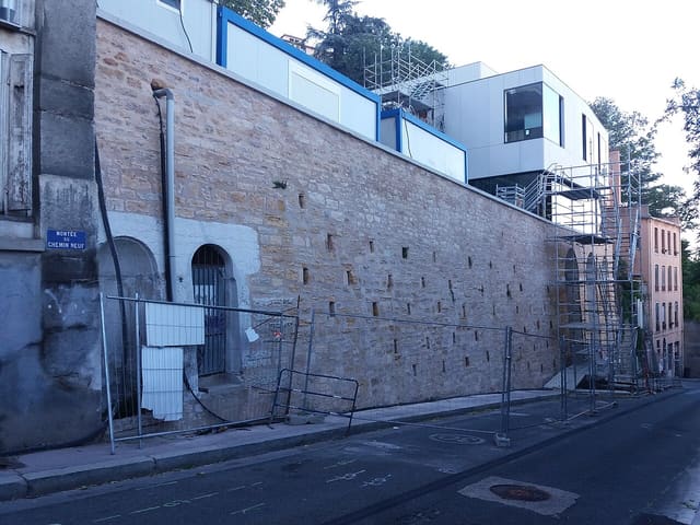 Le bas de la montée du Chemin-Neuf à Lyon, avec son haut mur de pierre, des barrières de chantier et la plaque de rue bleue.