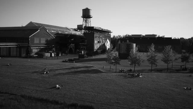 Parc aménagé sur une ancienne friche industrielle, avec promeneurs sur la pelouse devant d'anciens bâtiments, en noir et blanc