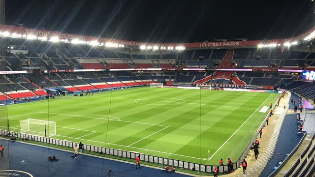 Le Parc des Princes de nuit, pelouse éclairée et tribunes vides avant un match, avec la bannière « Ici c'est Paris ».