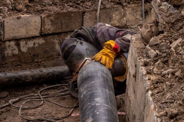A worker welds a pipe in an urban trench, showcasing industrial construction work.