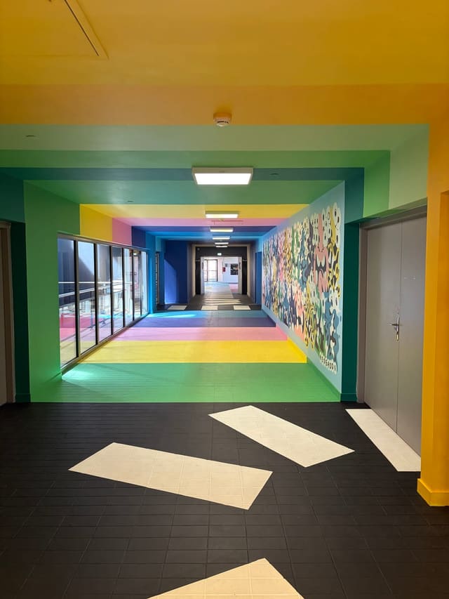 Un couloir de bureaux dont le sol, les murs et le plafond sont peints en bandes de couleurs vives ; sur la droite, une fresque de personnages enchevêtrés