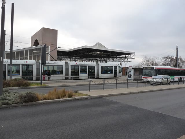 Une rame de tramway à l'arrêt devant la gare de Vénissieux, sous les lignes aériennes, avec un bus TCL à droite.